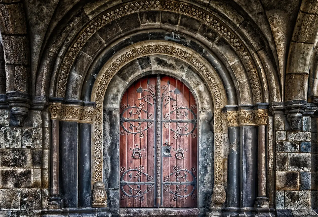 An ornate arched wooden door with a red finish and iron scrollwork, closed in a stone doorway